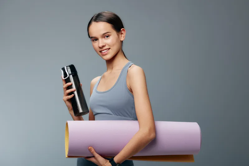 Fit woman in athletic tank stands with a water bottle and rolled yoga mat against a plain background, ready for a workout or fitness session.