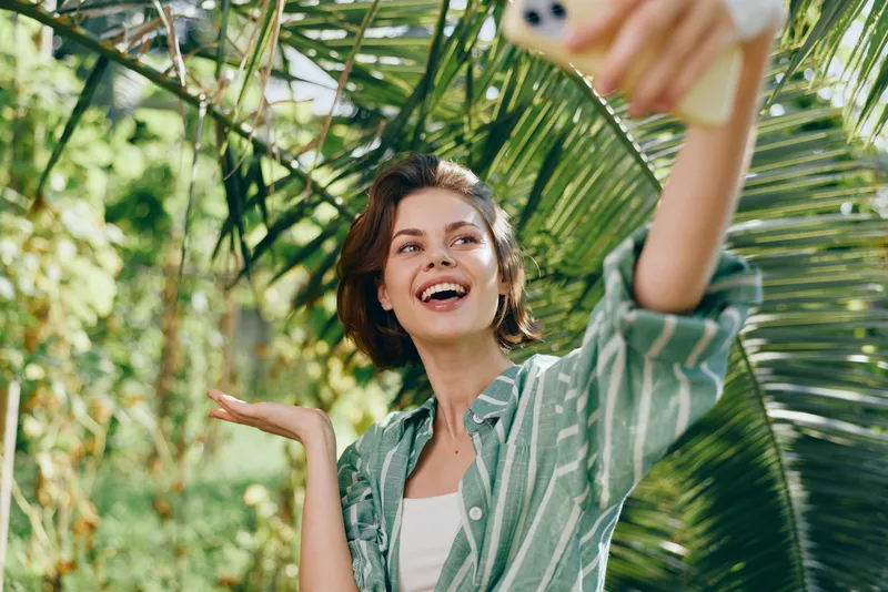 A joyful woman takes a selfie in a sunny outdoor setting amid lush greenery, wearing a striped shirt. She smiles broadly, capturing a carefree moment of happiness and confidence.