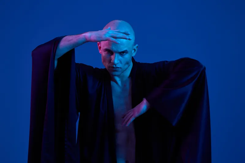 Man posing dramatically in blue lighting, showcasing artistic expression through body movement and attire. The scene emanates creativity and power with a captivating presence.