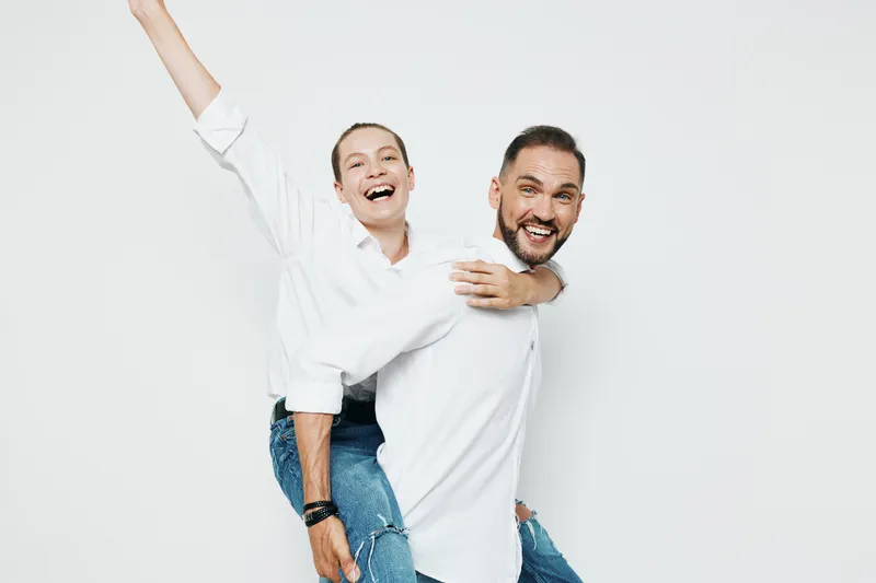 A professional man in business attire stands with a colleague on his back, arms wide in triumph against a clean white background, conveying confident teamwork and upbeat energy.