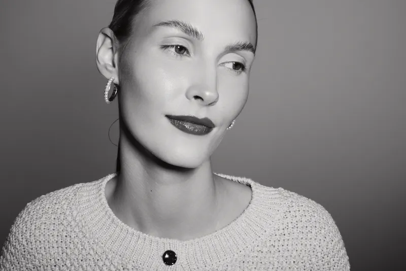 Portrait of a light skinned Caucasian woman with short hair wearing earrings and a knitted sweater, looking away with a soft smile in monochrome studio lighting.