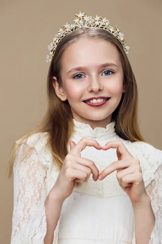 Young girl in elegant dress showing heart sign, smiling with a crown on her head against a soft beige background, conveying joy and innocence