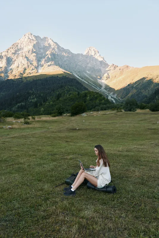 A young woman sits on a grassy meadow with a laptop, set against towering mountains. She concentrates on her screen, creating a calm, productive remote work moment outdoors.