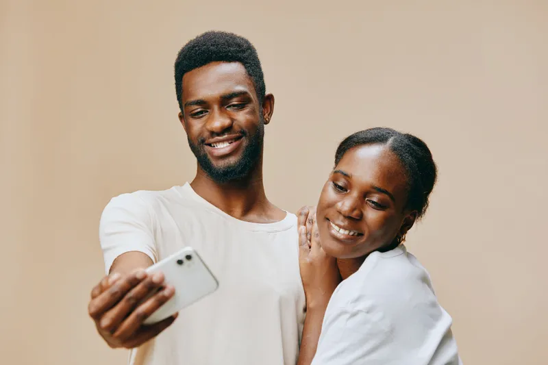 Young Black couple sharing a joyful moment while taking a selfie together against a soft beige background, showcasing love and happiness