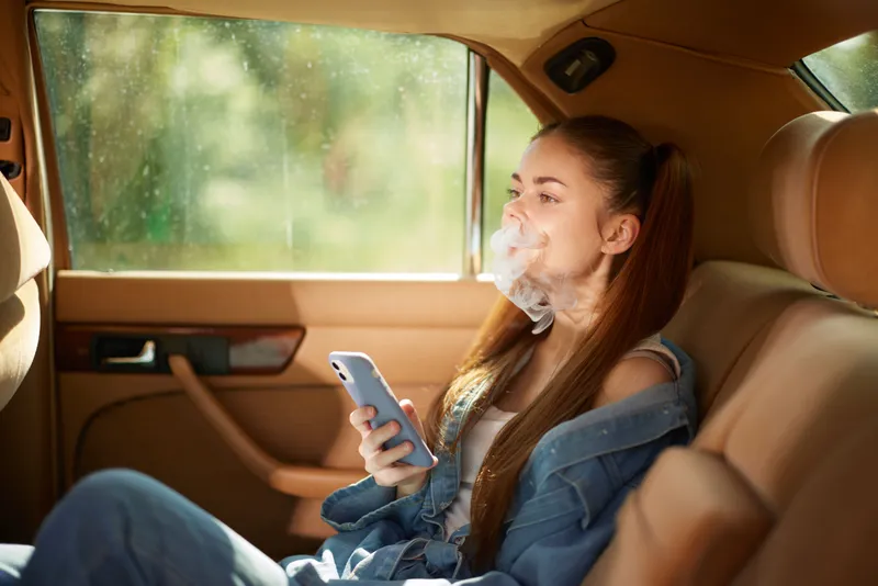 Young woman using smartphone in a car while exhaling smoke, dressed casually, reflecting a relaxed lifestyle in a sunny environment.