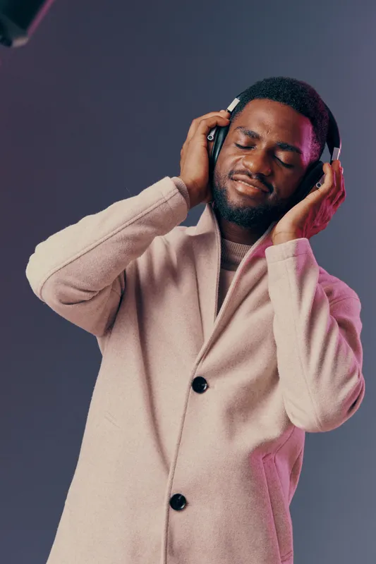 A joyful man enjoying music with closed eyes, wearing a stylish beige coat and headphones, surrounded by a soft gray background with vibrant lighting accents