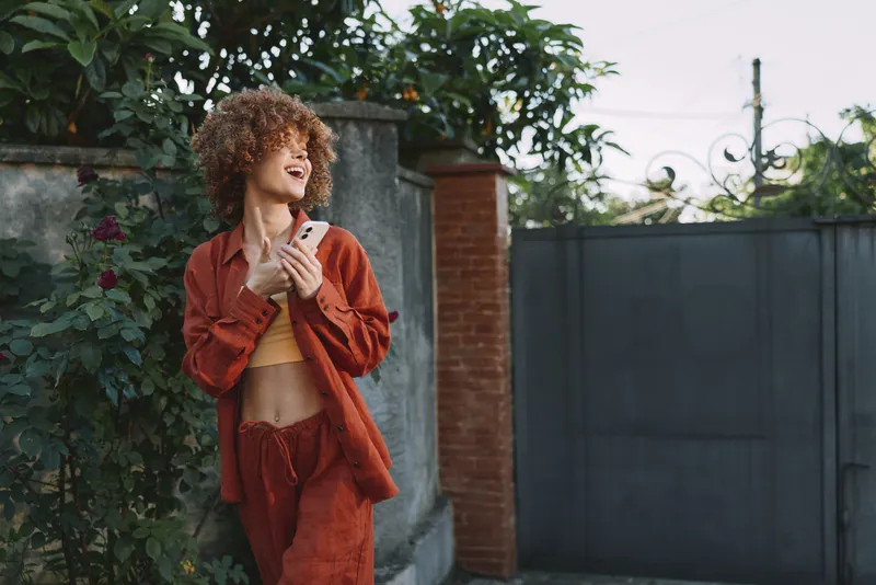 Woman outdoors in a rust-colored outfit, smiling with eyes closed, relaxed pose in sunlight, gentle breeze, lush greenery background, casual lifestyle moment, natural tones