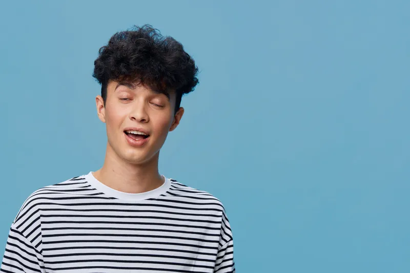 Smiling young man with closed eyes wearing a striped shirt against a soft blue background, expressing relaxed and joyful emotions with curly dark hair.