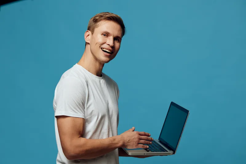 Smiling young man in casual white t-shirt holding a laptop computer, cheerful expression, isolated on solid blue background, studio portrait, people lifestyle concept, technology, communication, work,