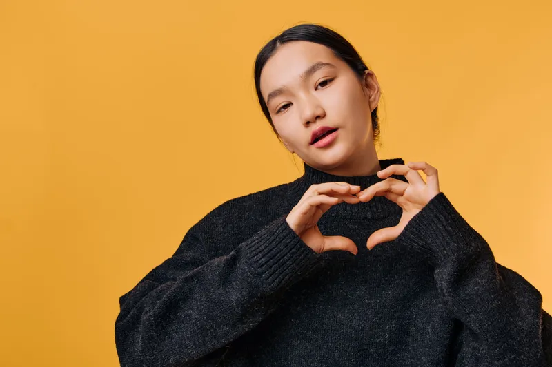 Young woman wearing black sweater forming heart shape with hands in front of orange background. Portrait of female showing love gesture with casual attire and natural expression.