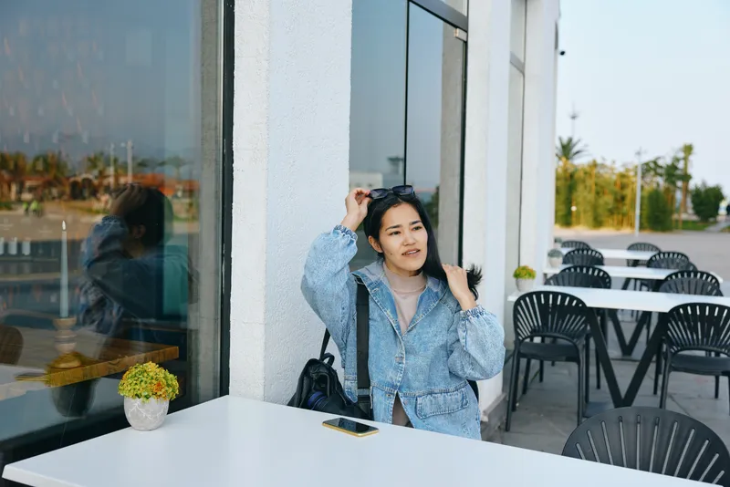 Cafe outdoor seating with a denim jacket woman at a white table, glass wall ambiance, phone on tabletop, plant centerpiece, casual street mood