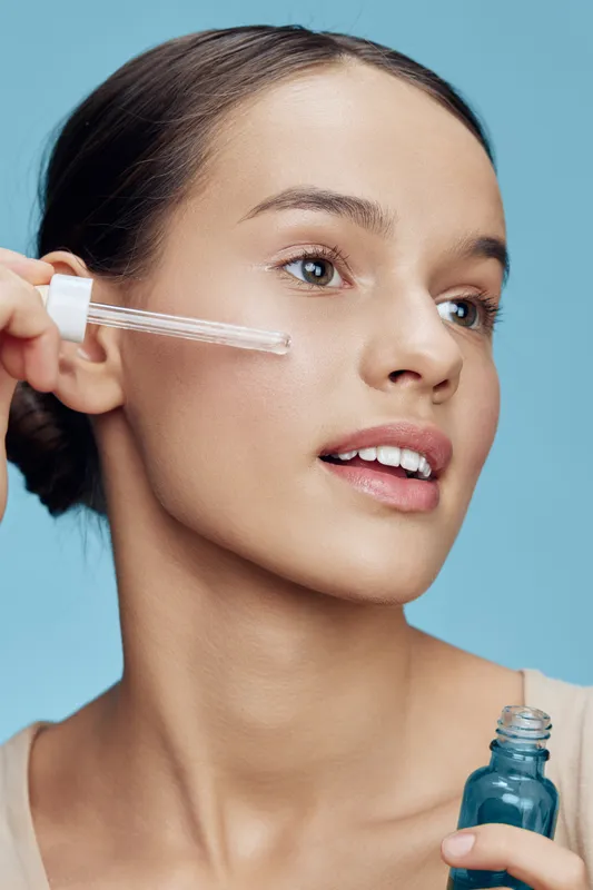 Young woman applying serum with dropper, showcasing radiant skin and a natural look against a soft blue background, emphasizing beauty and skincare