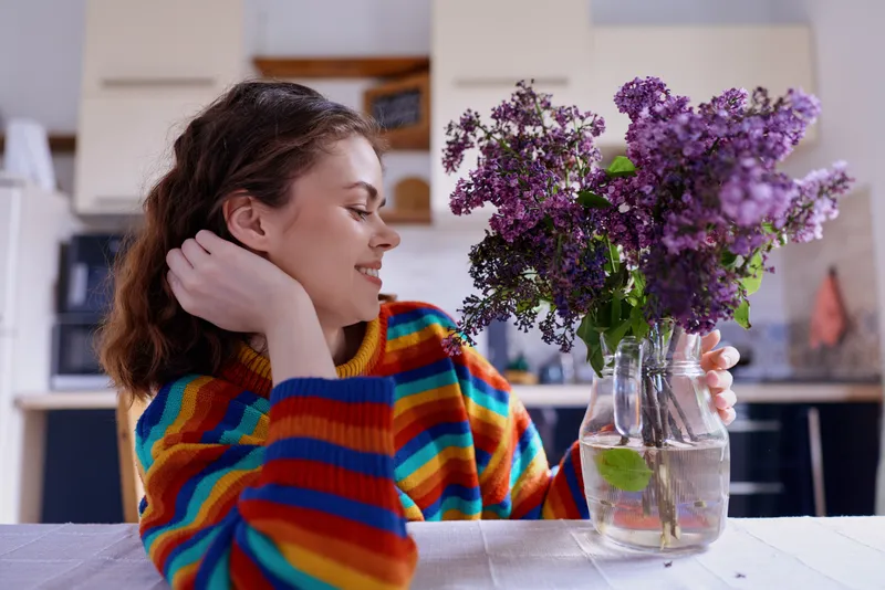 Woman at home sits beside a vase of lilac flowers on a table, smiling softly, wearing a striped sweater, creating a warm, cozy lifestyle moment in a bright kitchen setting and warm vibe