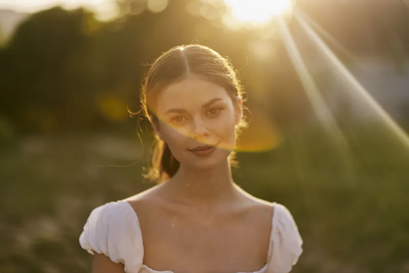A serene young woman in a flowing white dress poses gracefully at sunset, embodying tranquility, beauty, and nature in a vibrant outdoor setting
