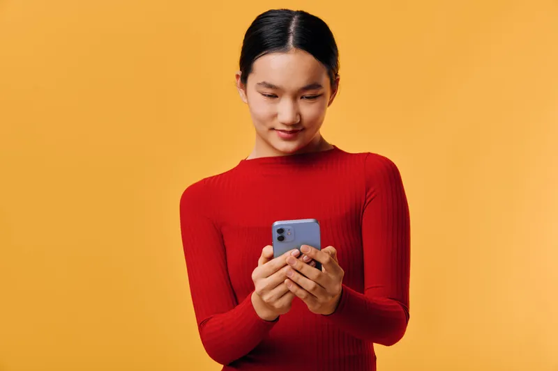 Young woman in red sweater holding smartphone and looking at screen with smile on orange background. Casual portrait, technology, communication and social media concept with digital device.