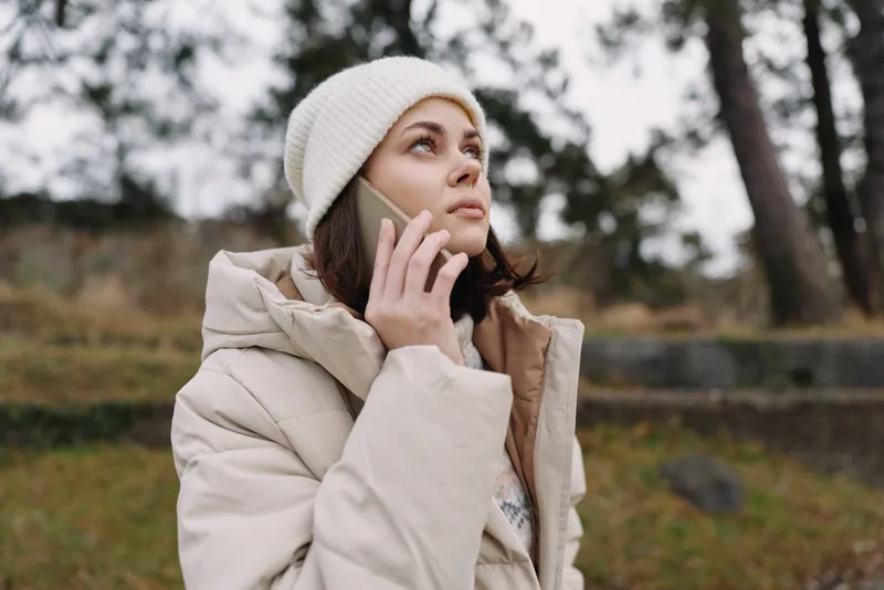 Woman talking on phone outdoors in a beige coat and beanie, standing in a park with bare trees, portraying a moment of mobile communication in chilly weather.