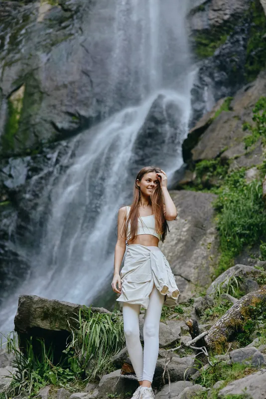 young woman standing near a waterfall in white outfit, enjoying nature, lush greenery, relaxed mood, outdoor adventure, scenic background, summer season