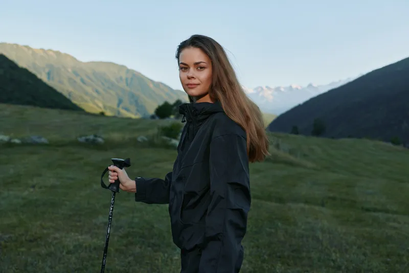 A lone hiker stands in a grassy mountain valley with a trekking pole. She gazes toward distant peaks, dressed in dark outdoor gear, projecting focus and determination.