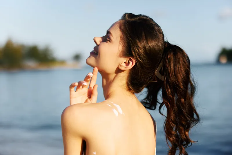 Portrait of a carefree woman by a calm lake, sunlight on skin, hair in a ponytail, sunscreen on shoulder, capturing warm summer mood and quiet seaside bliss.
