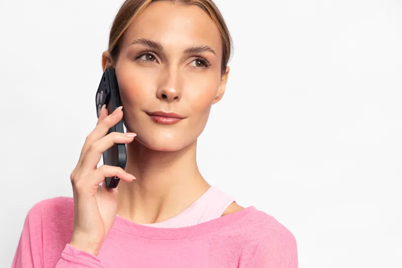 Young woman in pink sweater talking on smartphone, looking away with calm expression. Isolated on white background. Concept of communication, technology, and modern lifestyle.