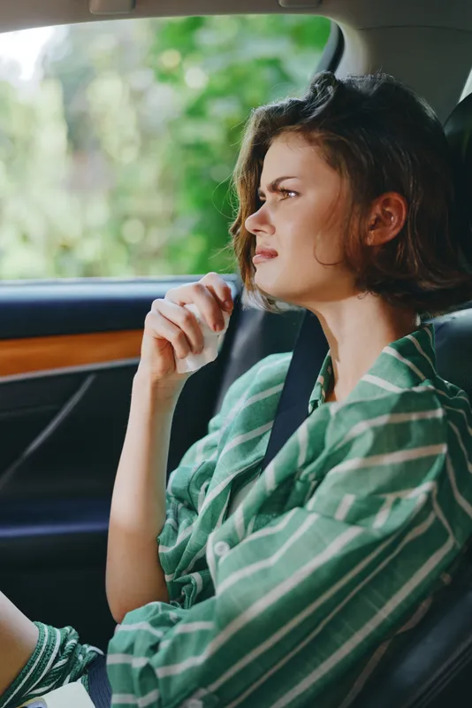 woman sits inside a car, looking ahead with calm thoughtful expression, hands near face, green striped shirt, soft light through window, car interior details, ready for a journey, peaceful moment