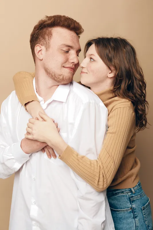 Young couple embraces tenderly, woman hugging man from behind with warm smile. Casual clothing and neutral background create intimate, loving, and affectionate mood in studio portrait.