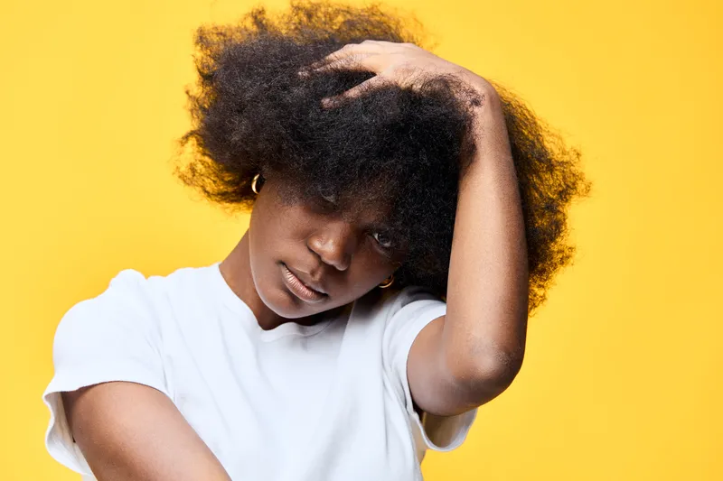Portrait of a young Black woman with curly hair, wearing a white shirt against a bright yellow background, expressing a confident and thoughtful emotion