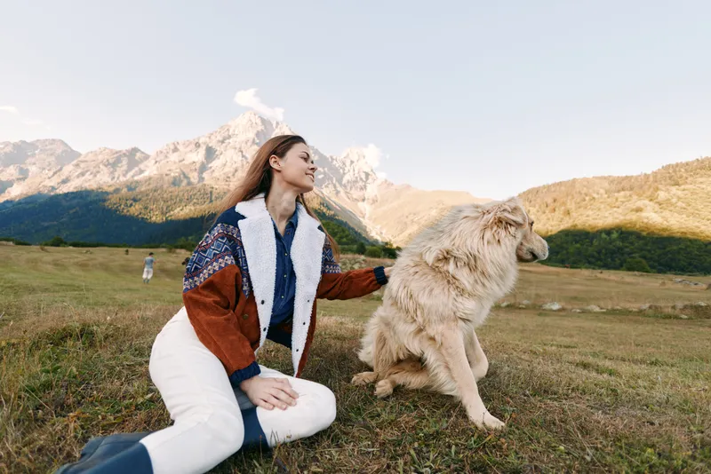 woman and dog sitting in a mountain meadow, nature scene with pet enjoying outdoors, relaxed portrait of female owner with fluffy dog, scenic landscape and clear sky