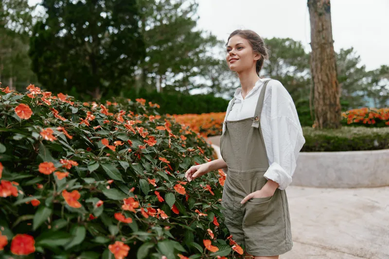 Young woman enjoying nature in overalls with flowers, smiling, in a serene park setting, reflecting a fresh and vibrant lifestyle