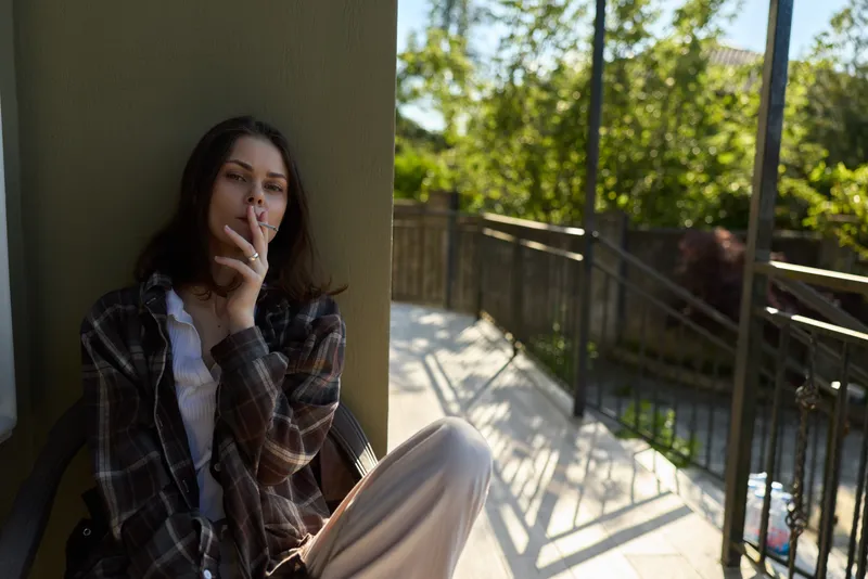 Casual lifestyle portrait of a woman smoking on a balcony, relaxed pose in daytime sunlight, plaid shirt, outdoor setting, calm and introspective mood