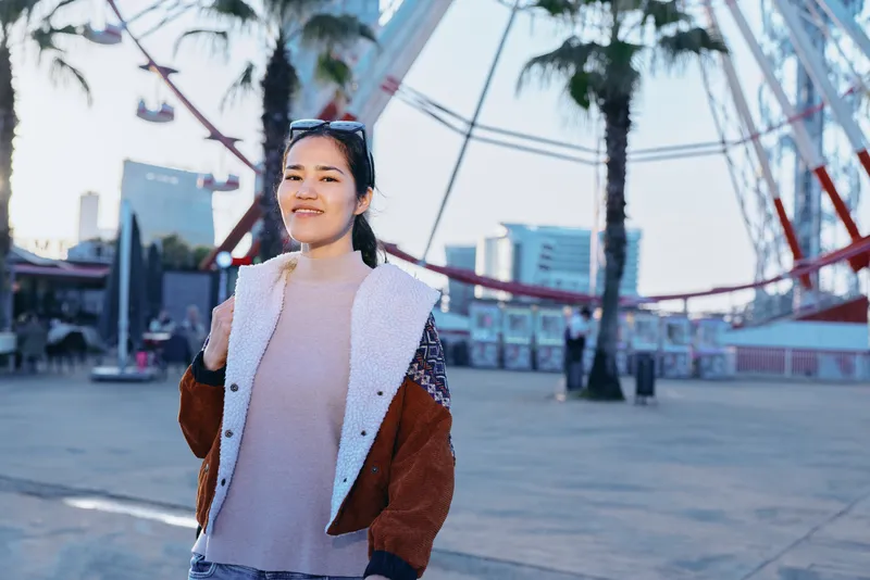 Woman wearing a cozy jacket in an outdoor urban setting near a modern bridge and palm trees, casual fashion and sunny mood captured in a wide public space
