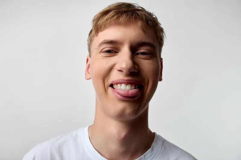 Smiling young man with tongue out, a playful expression and casual look in a studio shot with bright lighting against a white background, conveying fun, energy and youthful charm