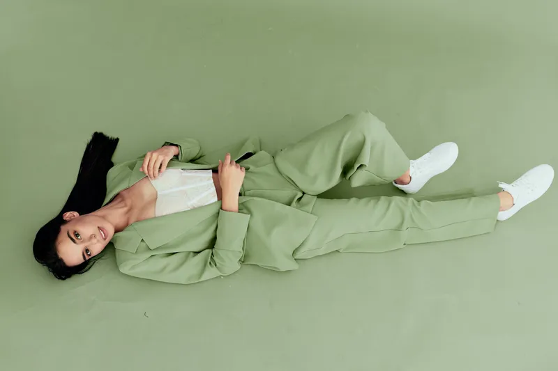 Woman lying on green floor wearing light green suit and white sneakers, relaxed pose, casual fashion, minimalistic background, modern style, soft natural lighting.