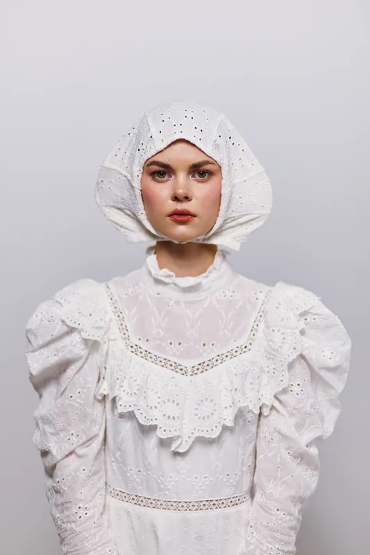 Young woman in white vintage dress and bonnet, expressing calmness and simplicity, against a soft gray background, ideal for seasonal themes and rustic concepts