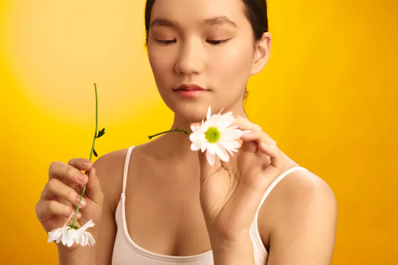 asian woman holds a white daisy close to her face, eyes gently closed, in a bright yellow studio, conveying calm focus and simple beauty and warmth.