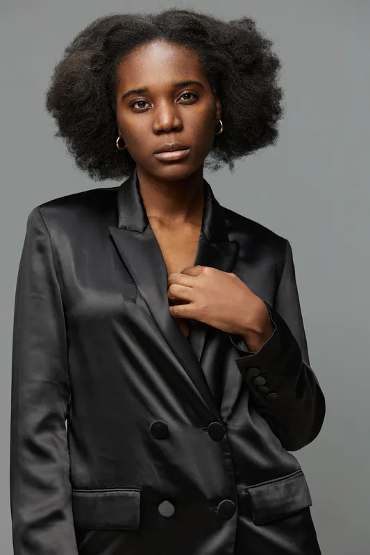 Confident young woman in a sleek black blazer, showcasing natural afro hair against a grey background, embodying modern elegance and empowerment