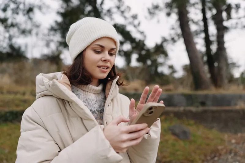 Smartphone, woman, outdoor, winter, jacket, beanie, cold weather interaction with device in hand in a park, focused on screen and calm surroundings