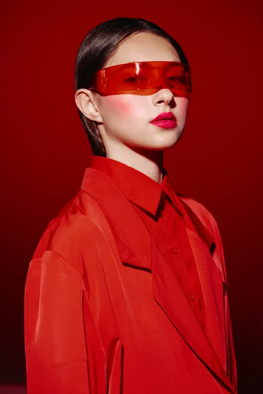 Portrait of a stylish young woman wearing bright red sunglasses and a matching oversized coat. She poses confidently against a vibrant red background. Concept for beauty and fashion creativity.
