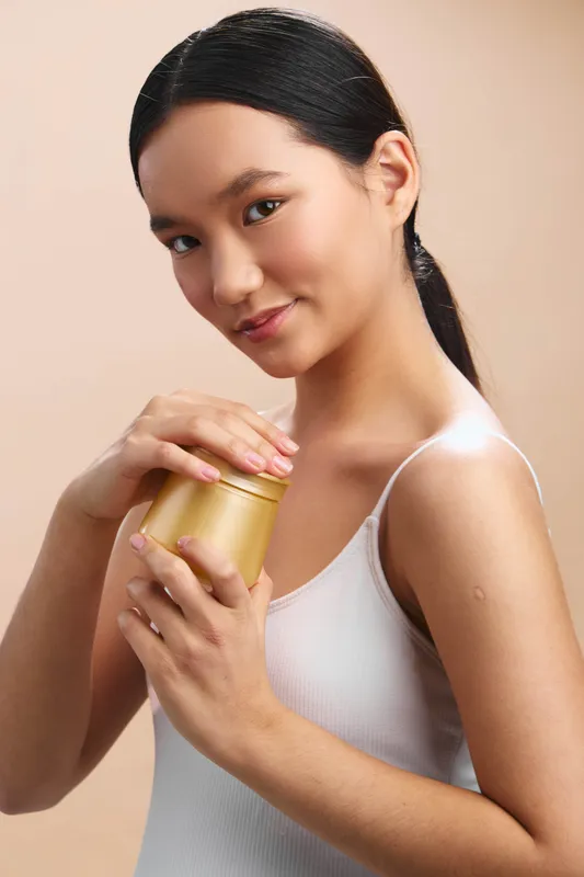 asian woman holds a gold jar close to her chest, smiling softly in a clean studio setting, presenting skincare product with calm confidence and warm lighting.