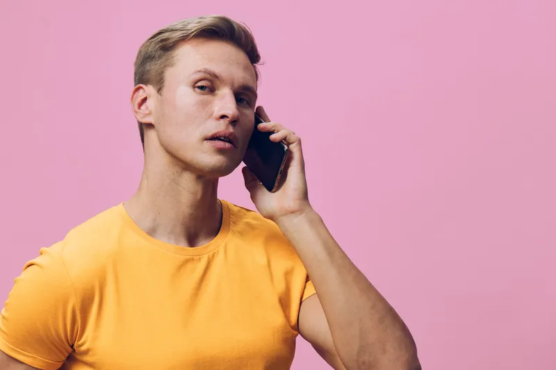 Man talking on mobile phone, casual yellow T-shirt, short hair, serious expression, vibrant pastel pink background, studio shot, modern communication, people lifestyle concept