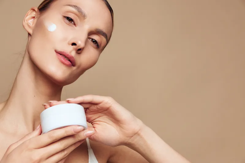 A young woman with clear skin applies white cream to her face while holding a jar of moisturizer. The neutral background highlights skincare and beauty care concepts.