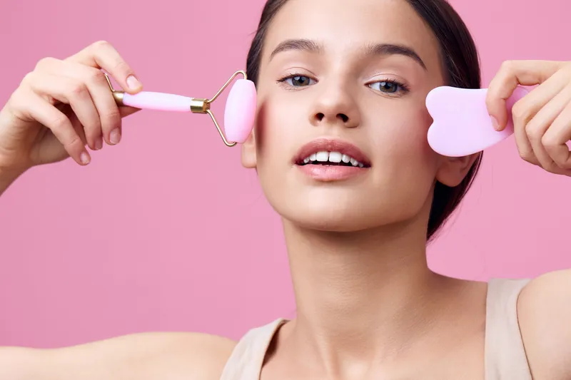 Young woman applying facial skincare with a pink roller and heart shaped tool on a pink background, promoting healthy skincare and beauty routines