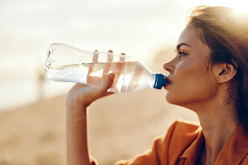 Woman drinking water from plastic bottle outdoors, close up side profile, calm focused expression, hydration and wellness concept, warm sunlight bokeh background, healthy lifestyle