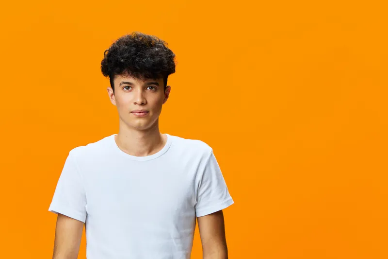 young man with curly hair wearing white t-shirt standing against bright orange background, casual style, confident expression, studio portrait lighting