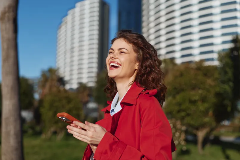 Young woman in a red jacket laughs outdoors in a sunny park with modern buildings in the background, holding a smartphone.