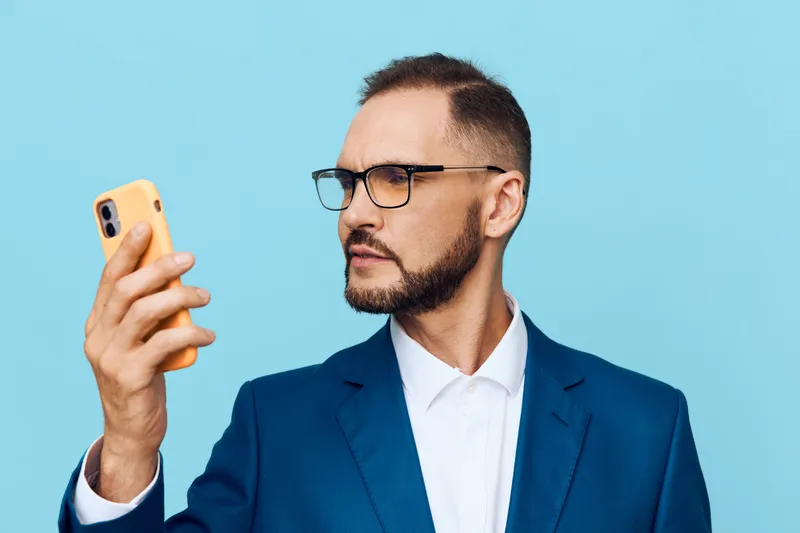 Professional businessman on a solid colored background, focused and confident, as he checks a mobile device. The isolated blue backdrop emphasizes ambition, clarity, and purpose in a modern work