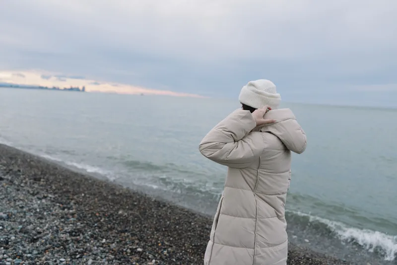 Person in a warm coat and knit hat stands on a pebbled beach facing a calm sea, wind brushing waves under a cloudy horizon for a serene seaside moment