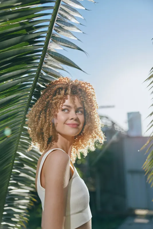 smiling woman with curly hair enjoys sunny outdoor setting near palm leaves, wearing white sleeveless top, fresh natural light and urban background