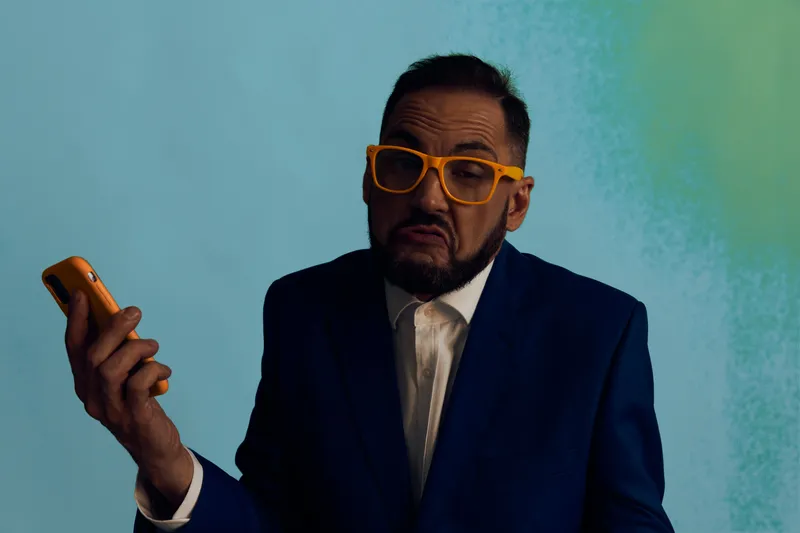 A business professional man stands against a colored isolated background, wearing a dark suit and bright orange glasses while checking his phone, conveying a focused, confident emotion.