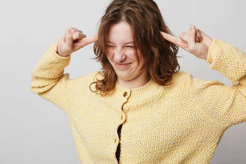 Young woman in yellow knitted sweater makes playful face and covers ears with fingers. Casual style, indoor portrait on light background. Concept of fun, mood, and personality expression.